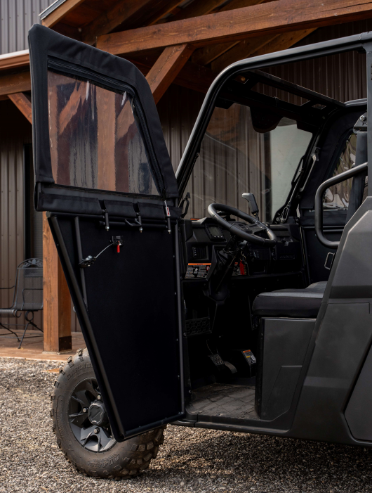 A Polaris Ranger 570SP Crew with Solid Convertible Doors is parked on gravel by a wooden structure, its driver’s door open to reveal a clear plastic window, soft cover, and the visible interior including steering wheel and dashboard.