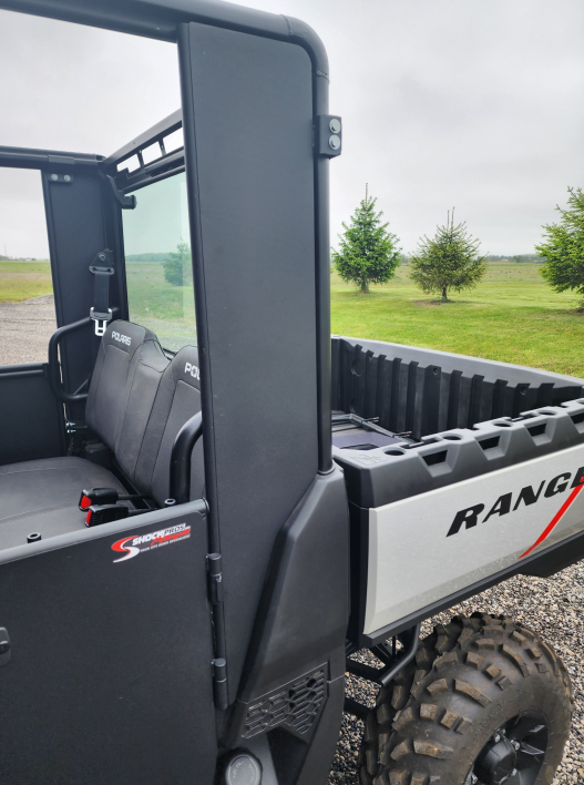 A close-up of a Polaris Ranger 570SP Crew with Solid Convertible Doors, featuring black seats and an empty cargo bed parked outdoors amid grass, evergreen trees, and a cloudy sky.
