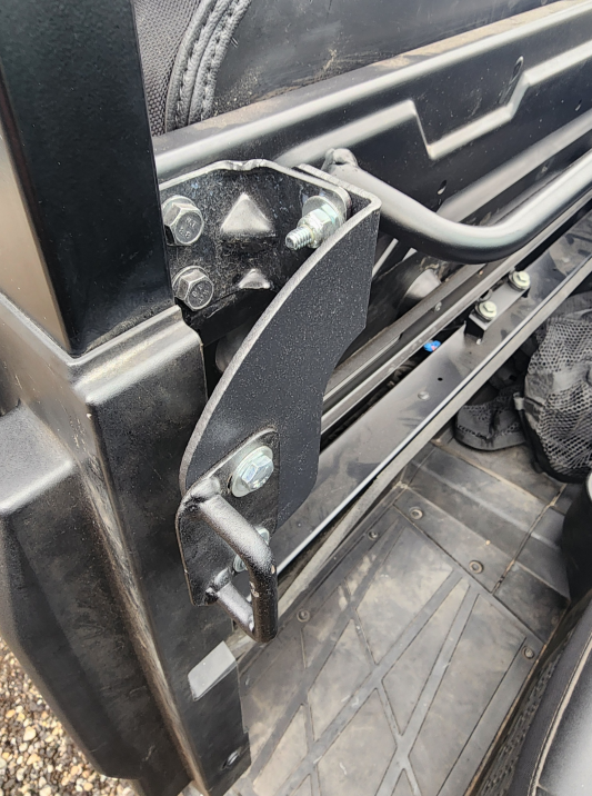 Close-up of a black metal hinge mechanism from the Polaris Ranger 570SP Crew Solid Convertible Doors, attached to the vehicle’s frame with large bolts near a textured rubber floor mat and mesh storage compartment. Part of the seat and frame are visible.