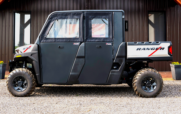 A Polaris Ranger 570SP Crew with solid convertible doors is parked on gravel before a brown metal building. The black utility vehicle has four doors, rugged tires, and a truck bed featuring a white RANGER logo with red accents.