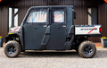 A Polaris Ranger 570SP Crew with solid convertible doors is parked on gravel before a brown metal building. The black utility vehicle has four doors, rugged tires, and a truck bed featuring a white RANGER logo with red accents.
