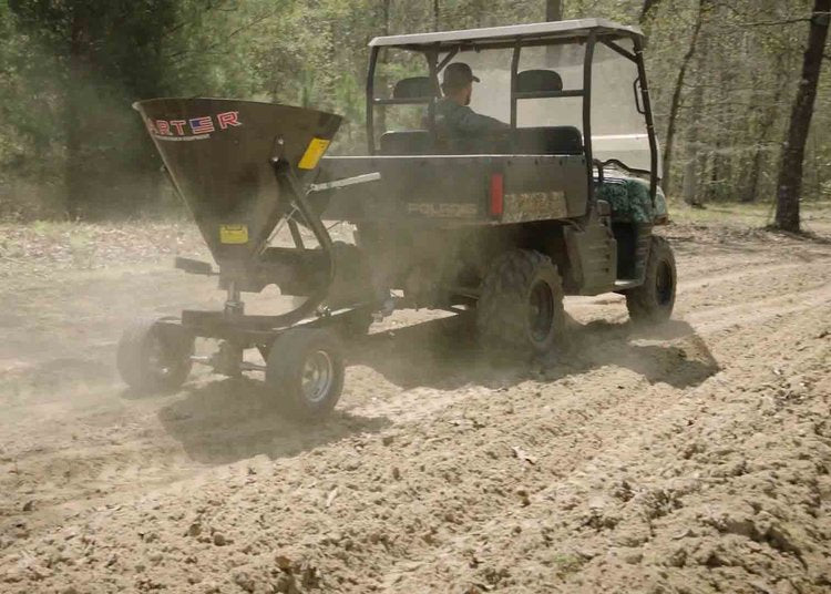 A utility vehicle tows the Heavy Duty Commercial Grade 5-Bushel Pull-Behind Spreader/Trail Feeder along a dirt path in a sunlit wooded area, dispersing material onto the soil and creating dust clouds behind it.