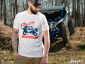 A bearded man in a white SuperATV Red, White, and Blue Unisex T-Shirt and black cap stands outdoors, looking down. A blue off-road vehicle sits in the wooded background. Tattoos cover his left arm; the SuperATV logo is in the bottom right corner.