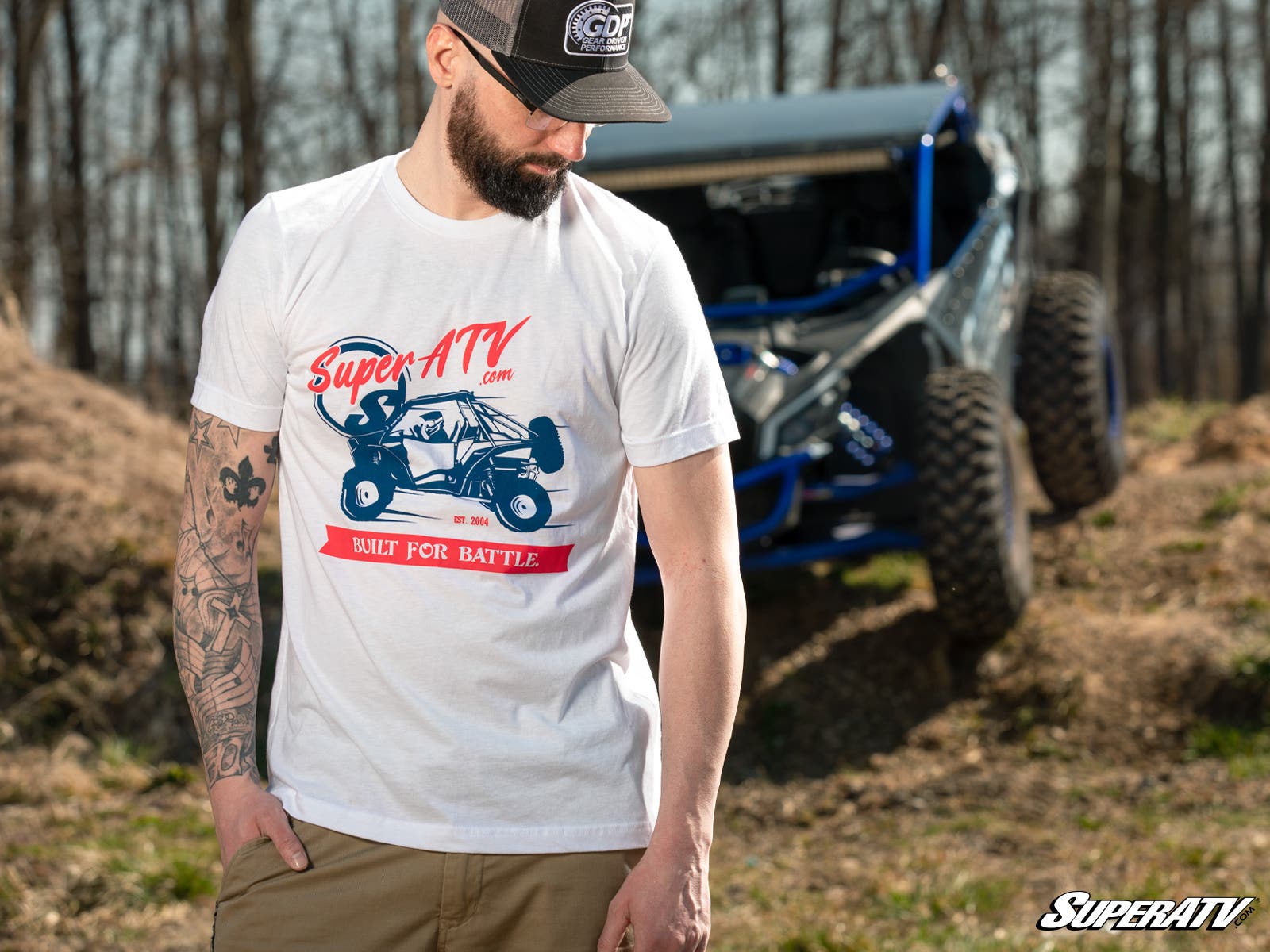 A bearded man in a white SuperATV Red, White, and Blue Unisex T-Shirt and black cap stands outdoors, looking down. A blue off-road vehicle sits in the wooded background. Tattoos cover his left arm; the SuperATV logo is in the bottom right corner.