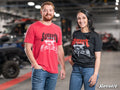 A man in a red and a woman in a black SuperATV Rock n Roll Unisex T-Shirt stand indoors, smiling, with off-road vehicles blurred in the background. The SuperATV logo appears at the bottom right.