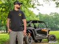A bearded man in sunglasses, a black cap, gray shorts, and the Man, Myth, Machine Unisex T-Shirt stands outdoors with hands in pockets. Behind him is an orange and black RZR off-road vehicle. The SuperATV logo is visible among green trees under a sunny sk