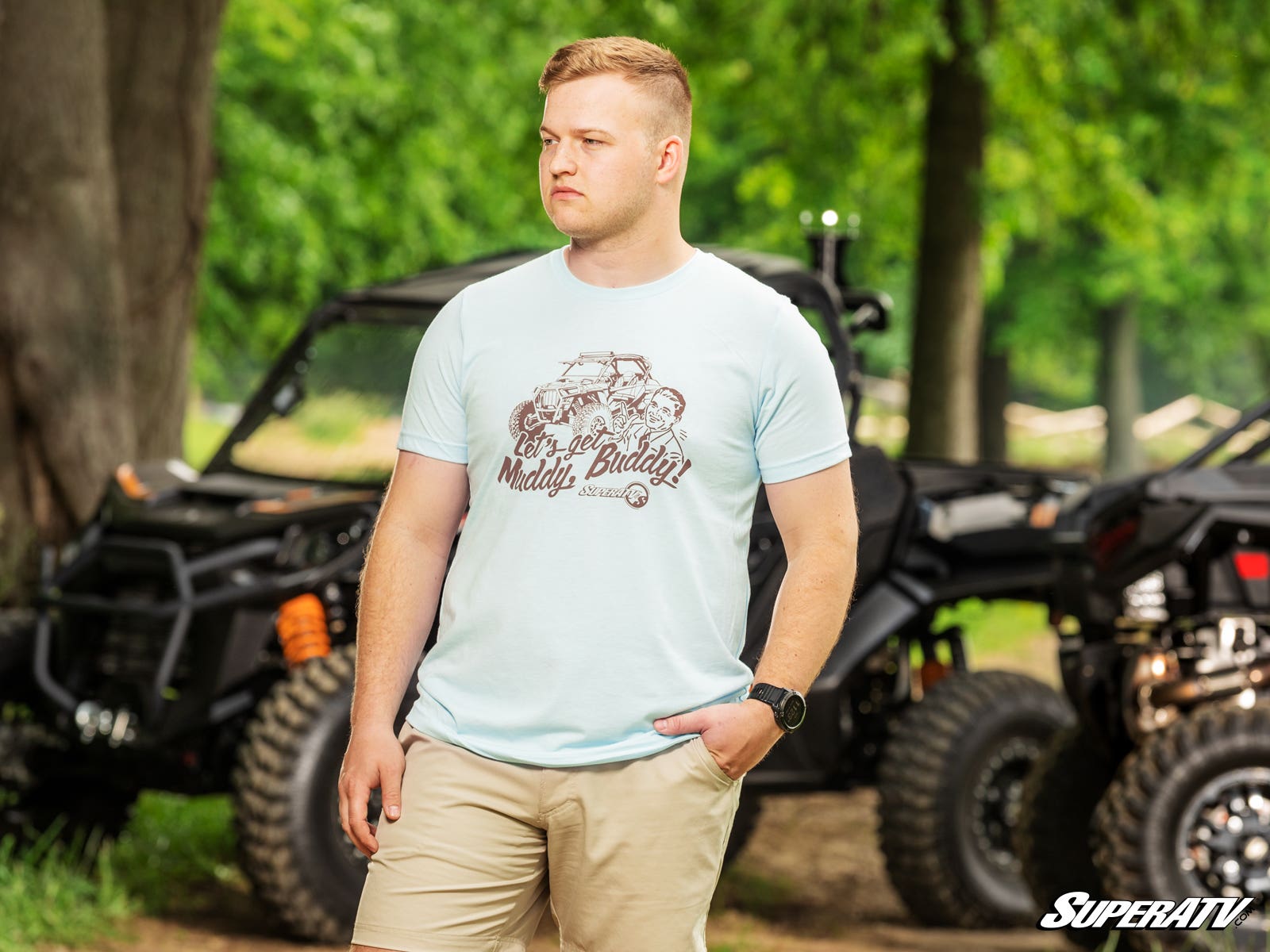 A young man in a Muddy Buddy Heather Ice Blue Unisex T-Shirt and khaki shorts stands outdoors near black off-road vehicles, looking to the side with a neutral expression amid trees and greenery. The “SuperATV” logo appears at the bottom right.