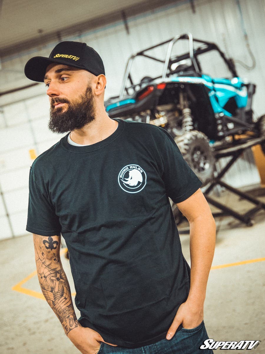 A bearded man in a Rhino Axles T-Shirt and black cap stands indoors, showing his tattoo sleeve. Behind him, a teal and black off-road vehicle is lifted on a stand in a workshop with metal walls. The image features the SuperATV brand.