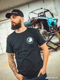 A bearded man in a Rhino Axles T-Shirt and black cap stands indoors, showing his tattoo sleeve. Behind him, a teal and black off-road vehicle is lifted on a stand in a workshop with metal walls. The image features the SuperATV brand.