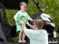 A smiling young girl in a green Mud Monster Kids T-Shirt stands on an ATV step, laughing as a tattooed adult helps steady her. The black and white Defender ATV is surrounded by green trees, with the SuperATV logo visible in the corner.