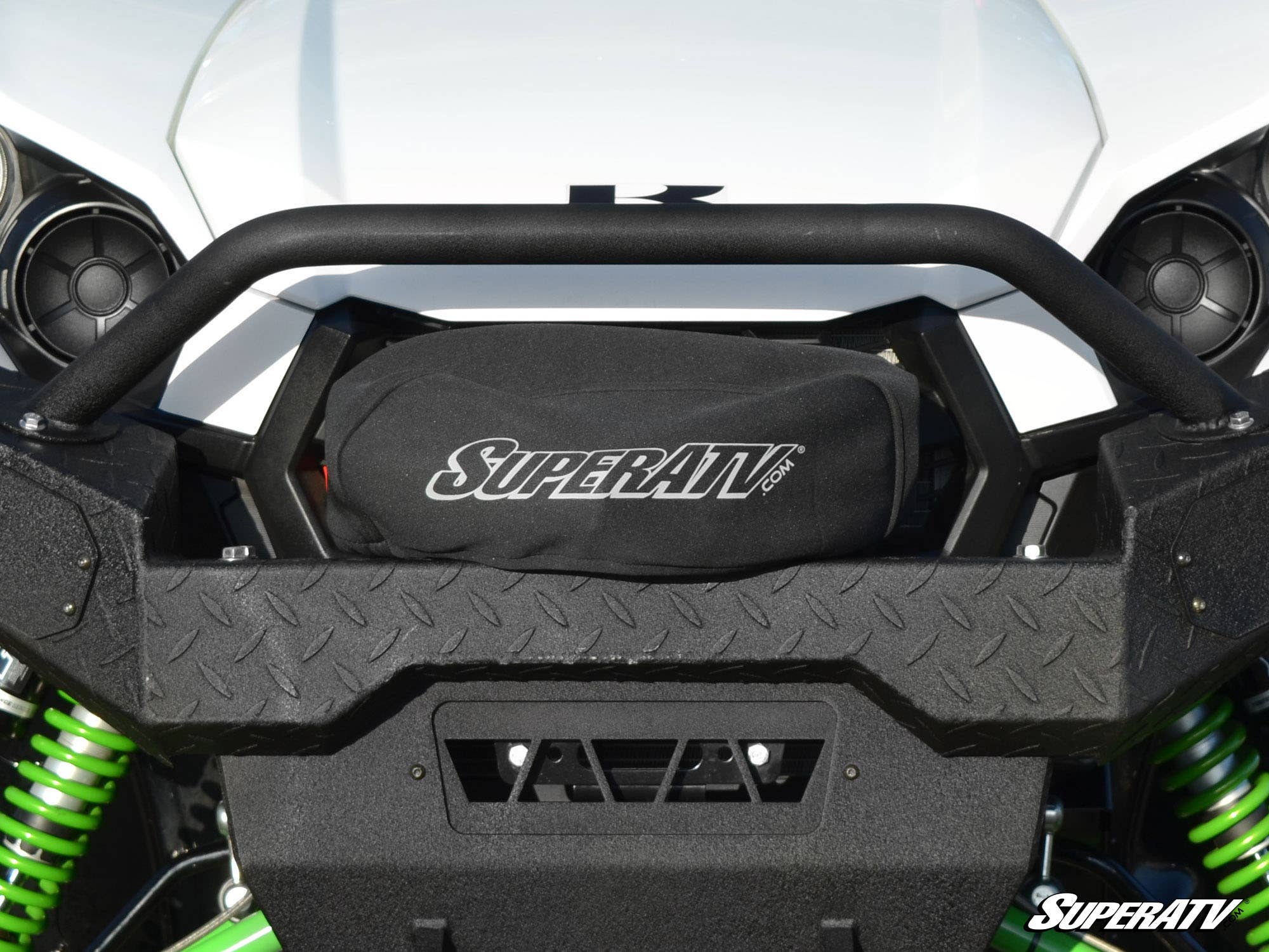 Close-up of a white off-road vehicle’s front bumper with a black SuperATV Winch Cover featuring the “SuperATV.com” logo, rugged diamond-plate texture, visible green suspension springs, and part of the “SuperATV” logo in the corner.