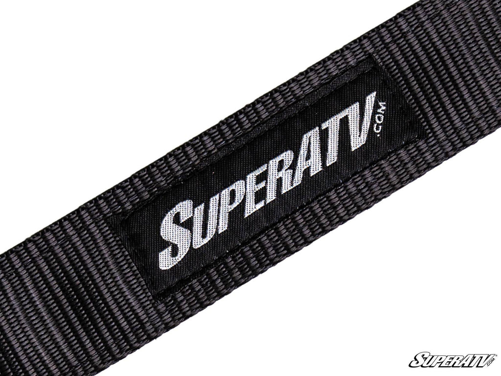 A close-up of a SuperATV Limit Strap shows a black woven strap with a rectangular black patch displaying “SUPERATV.COM” in white. The patch is sewn onto the strap, and a small SuperATV logo appears in the bottom right corner.