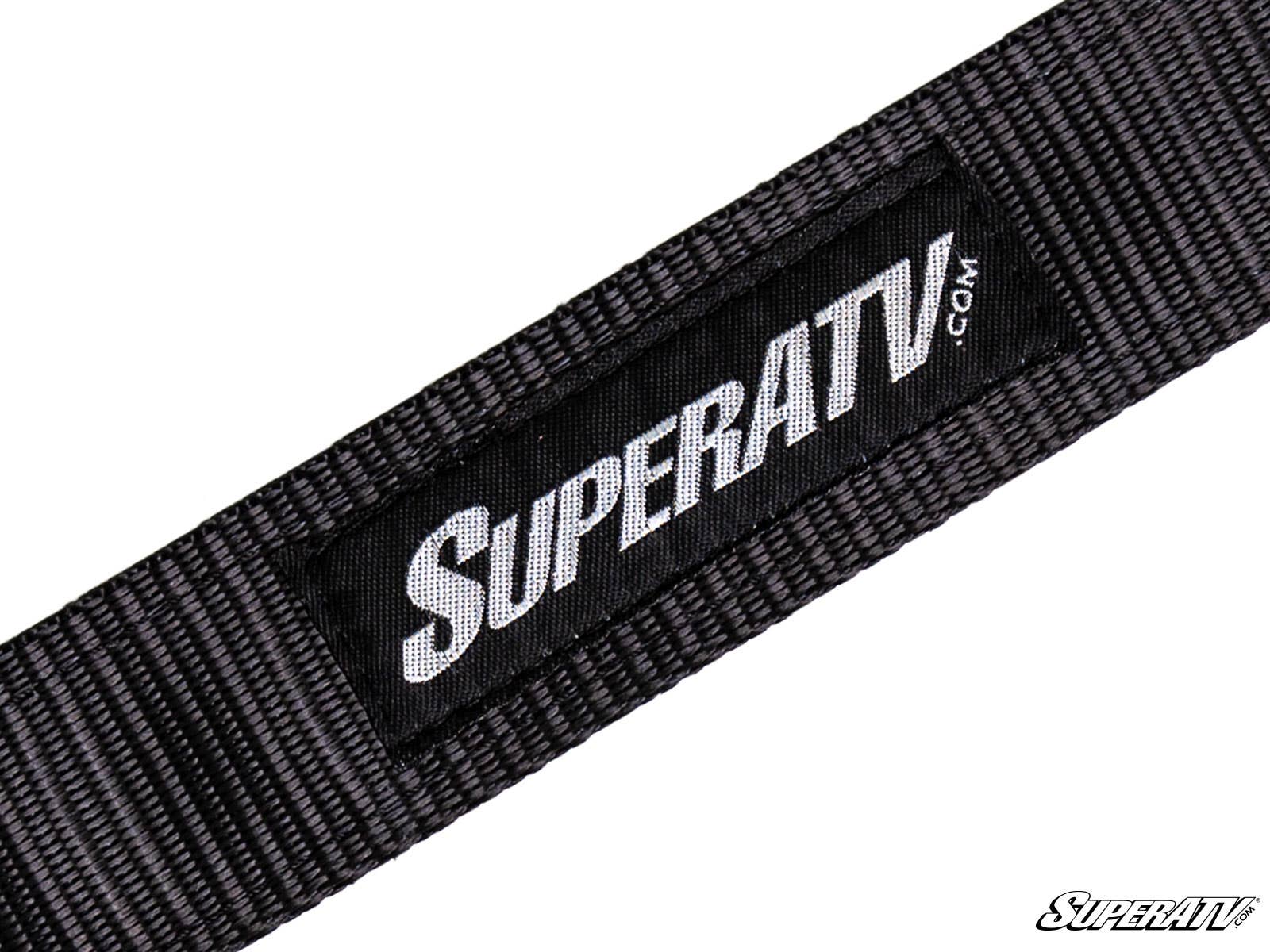 A close-up of a SuperATV Limit Strap shows a black woven strap with a rectangular black patch displaying “SUPERATV.COM” in white. The patch is sewn onto the strap, and a small SuperATV logo appears in the bottom right corner.