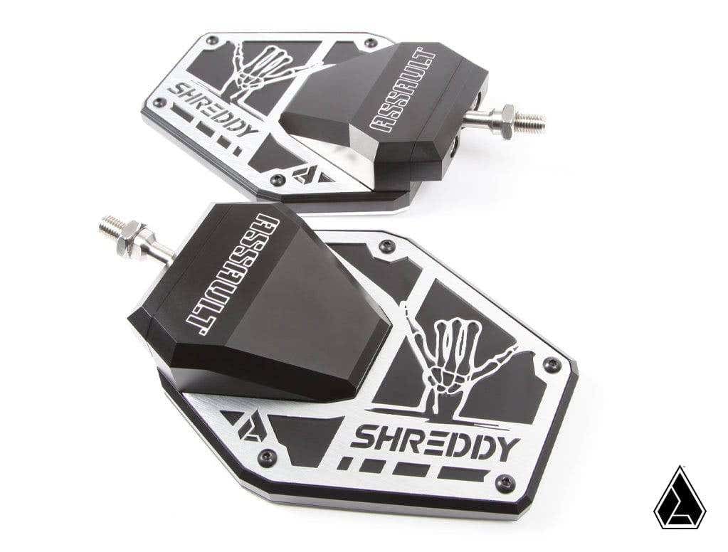 Two Assault Industries SHREDDY B2C Convex Side Mirrors with black angular mounts, silver bases featuring a white skeleton hand graphic, geometric cutouts, and mounting bolts. A small black triangle logo appears in the bottom right corner.