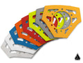 Six pentagon-shaped Assault Industries Steering Wheel Backing Plates in white, blue, red, green, silver, and orange are stacked and fanned out. Each has multiple holes and cutouts with a small black triangle logo at the bottom right on white.