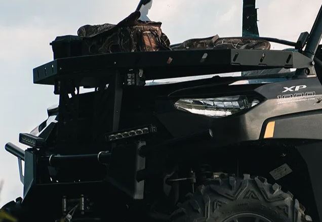 Close-up of a black 2014+ Polaris Ranger 900/1000/1000Xp with rugged tires and a Swampox Hood Rack mounted in front, carrying camouflaged gear. The XP 1000 branding is near the headlight, with sky and outdoor scenery in the background. Made in the U.S.A.