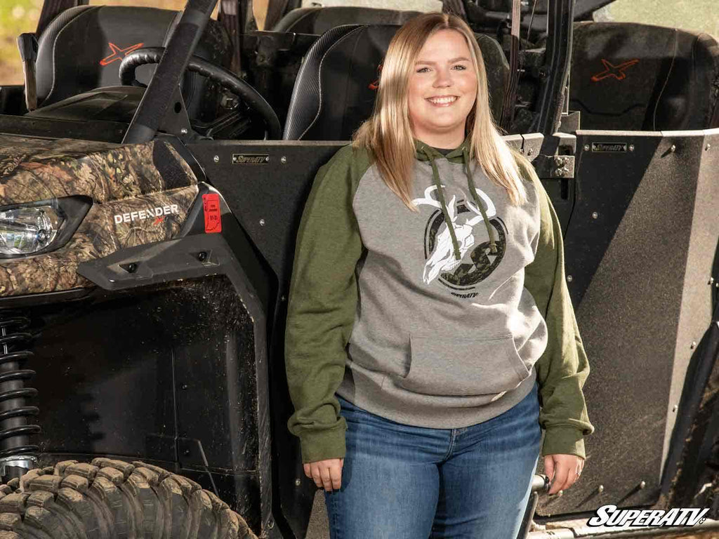 A smiling woman with straight blonde hair stands by a camo off-road vehicle, wearing the SuperATV Green and Gray Skull Hoodie and blue jeans. The seats, tires, and a “Super ATV” logo in the lower right corner are visible.