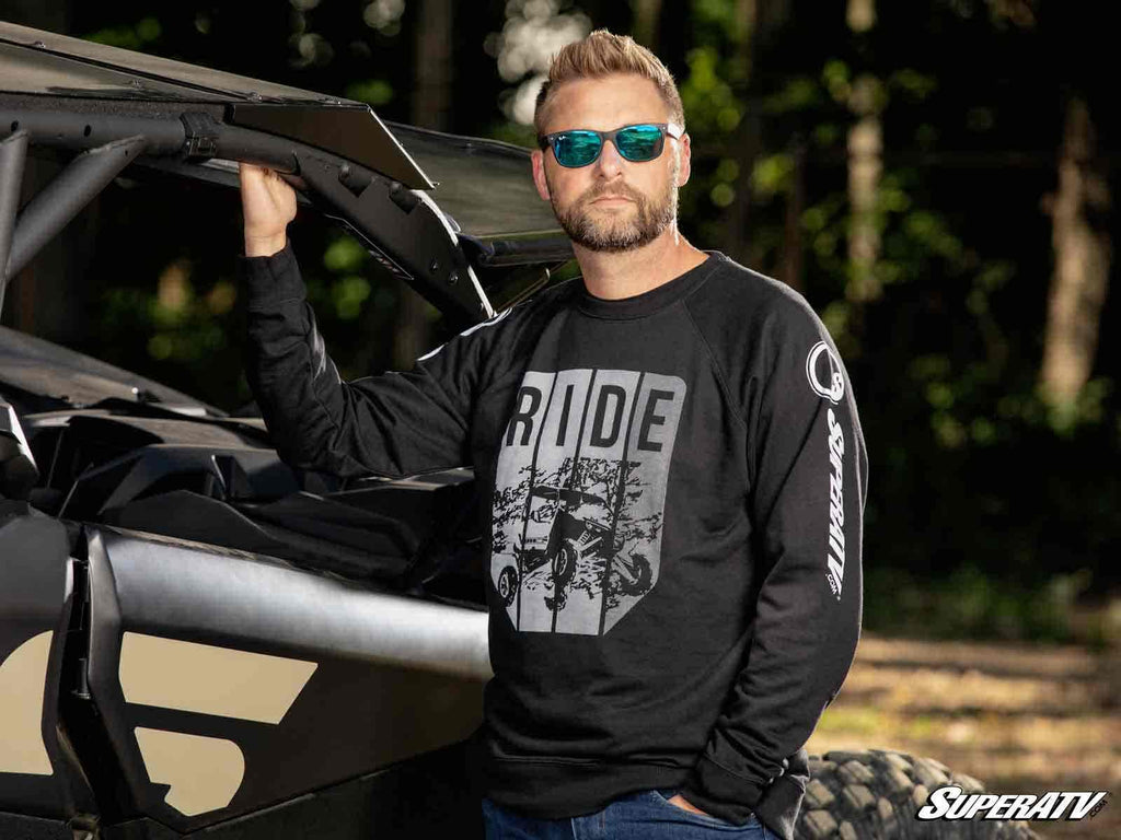 A man with a beard and sunglasses stands beside an off-road vehicle with the hood up, wearing a SuperATV Black Ride Sweatshirt. One hand rests on the vehicle as trees and sunlight blur in the background.