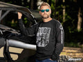 A man with a beard and sunglasses stands beside an off-road vehicle with the hood up, wearing a SuperATV Black Ride Sweatshirt. One hand rests on the vehicle as trees and sunlight blur in the background.