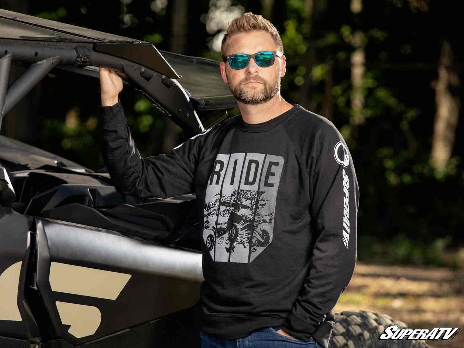 A man with a beard and sunglasses stands beside an off-road vehicle with the hood up, wearing a SuperATV Black Ride Sweatshirt. One hand rests on the vehicle as trees and sunlight blur in the background.