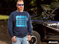 A bearded man with sunglasses and light hair stands outdoors in blue jeans, wearing a SuperATV Indigo Crewneck Sweatshirt beside a black off-road vehicle with blue and white details. The SUPERATV logo is in the bottom right corner.