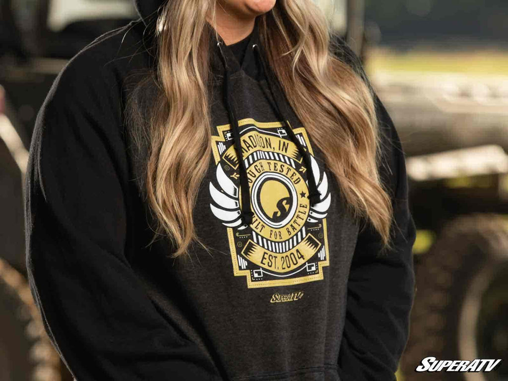 Someone with long, wavy blonde hair wears the SuperATV Black Built for Battle Hoodie with a yellow and white “Madison, IN—Winchester—Built for Battle—Est. 2004” graphic and SuperATV logo. The outdoor background is blurred.