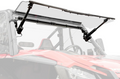 A close-up of a Honda Talon 1000 Max-Drive Power Flip Windshield, shown clear and upward-tilted with black brackets. The vehicles frame, steering wheel, and seat are visible beneath the partially raised windshield against a white background.