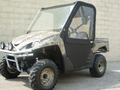 A Kawasaki Teryx with Convertible Doors and camouflage pattern, large rugged tires, clear windshield, black fabric doors, and a sturdy metal front bumper is parked on pavement in front of a beige cinder block wall.