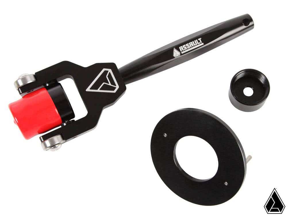 The Assault Industries RZR Secondary Belt Replacement Tool for select Polaris RZR Turbo models is shown in black and red with geometric logos, alongside two round black components, all made of metal and plastic on a white background.