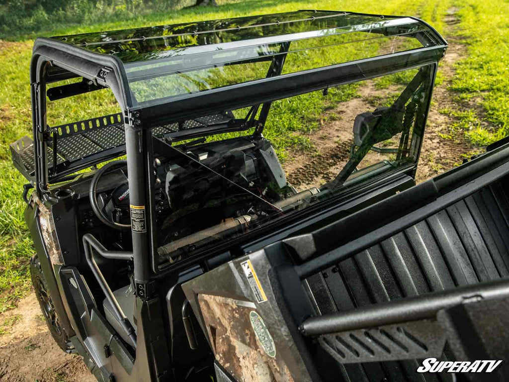 A Tracker 800SX with a clear glass windshield and roof is parked on a dirt path next to grass. The open cargo bed and steering wheel are visible. The SuperATV logo appears in the bottom right. Shown with Tracker 800SX Rear Windshield.