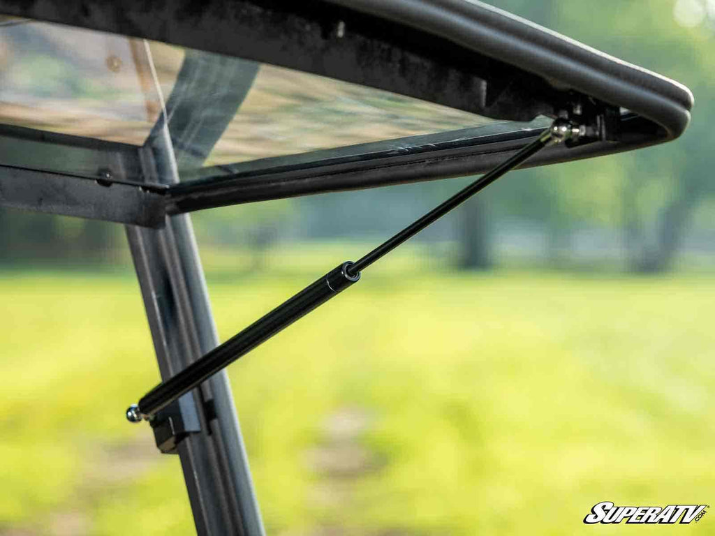 Close-up of a black hydraulic strut supporting the Tracker 800SX Flip Windshield on an off-road vehicle, connecting the frame and window to hold it open. Blurred grass and trees are in the background. The SuperATV logo appears in the bottom right corner.