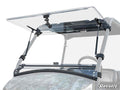 A Tracker OX400 Flip Windshield, made of clear polycarbonate, is partially open on a grey off-road vehicle with metal brackets and gas struts. The hinge mechanism is visible and the SuperATV logo is in the lower right corner.