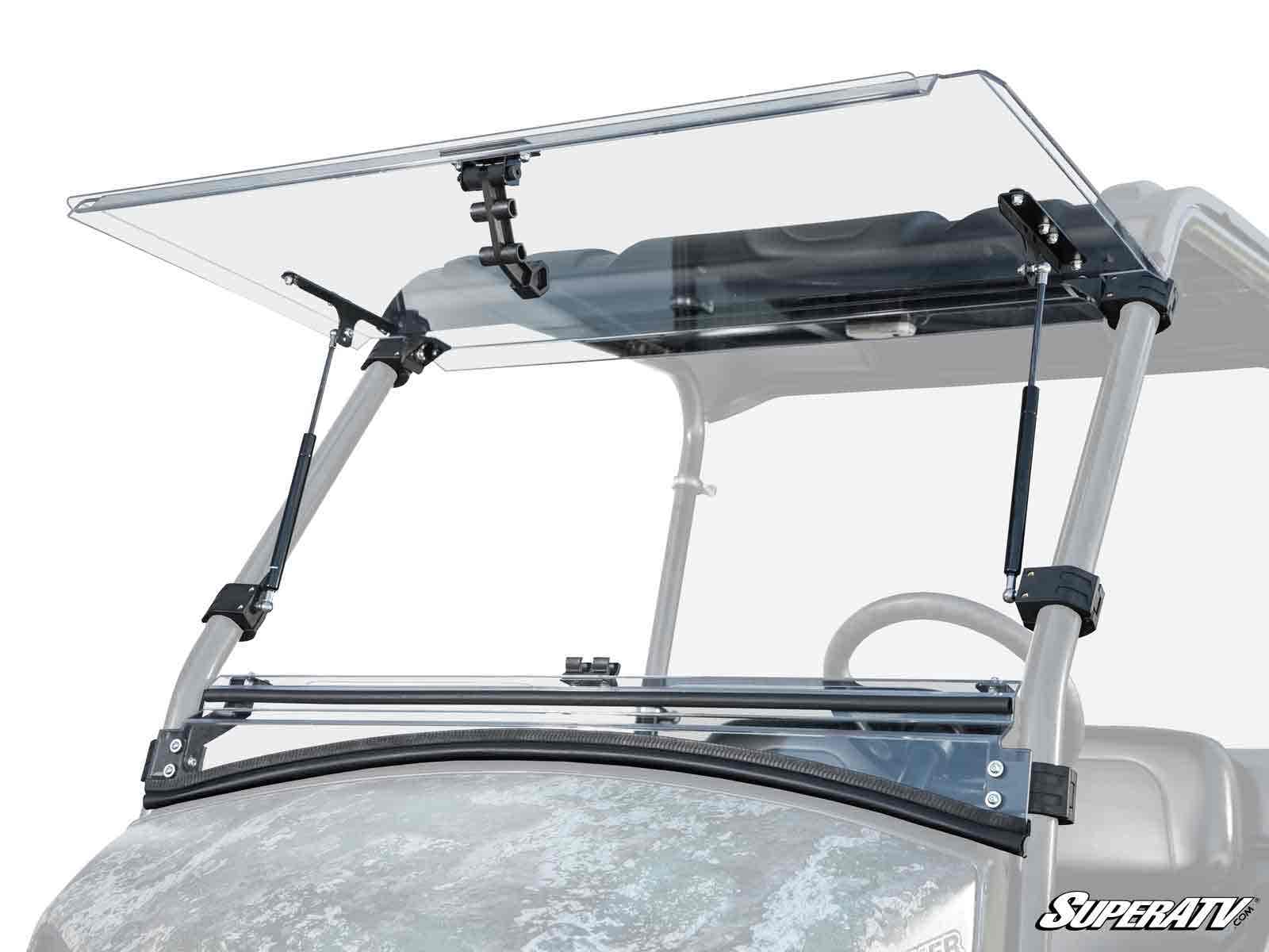 A Tracker OX400 Flip Windshield, made of clear polycarbonate, is partially open on a grey off-road vehicle with metal brackets and gas struts. The hinge mechanism is visible and the SuperATV logo is in the lower right corner.