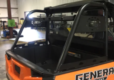 An orange Polaris General 1000 with a black roll cage is parked indoors. Its empty bed and background workshop equipment are visible. The word GENERAL appears partially on the side. Shown: Trail Armor Polaris General 1000 Rear Rack.