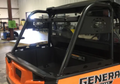 An orange Polaris General 1000 with a black roll cage is parked indoors. Its empty bed and background workshop equipment are visible. The word GENERAL appears partially on the side. Shown: Trail Armor Polaris General 1000 Rear Rack.
