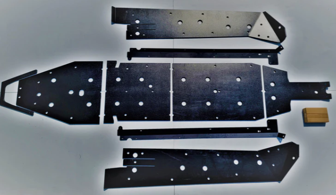 Five flat, black metal plates and two long bars from the Trail Armor Polaris Turbo R 4 Full Skids with Standard or Trimmed Sliders are arranged on a white surface, with a small brown box nearby for scale; the plates feature multiple holes and angular edge
