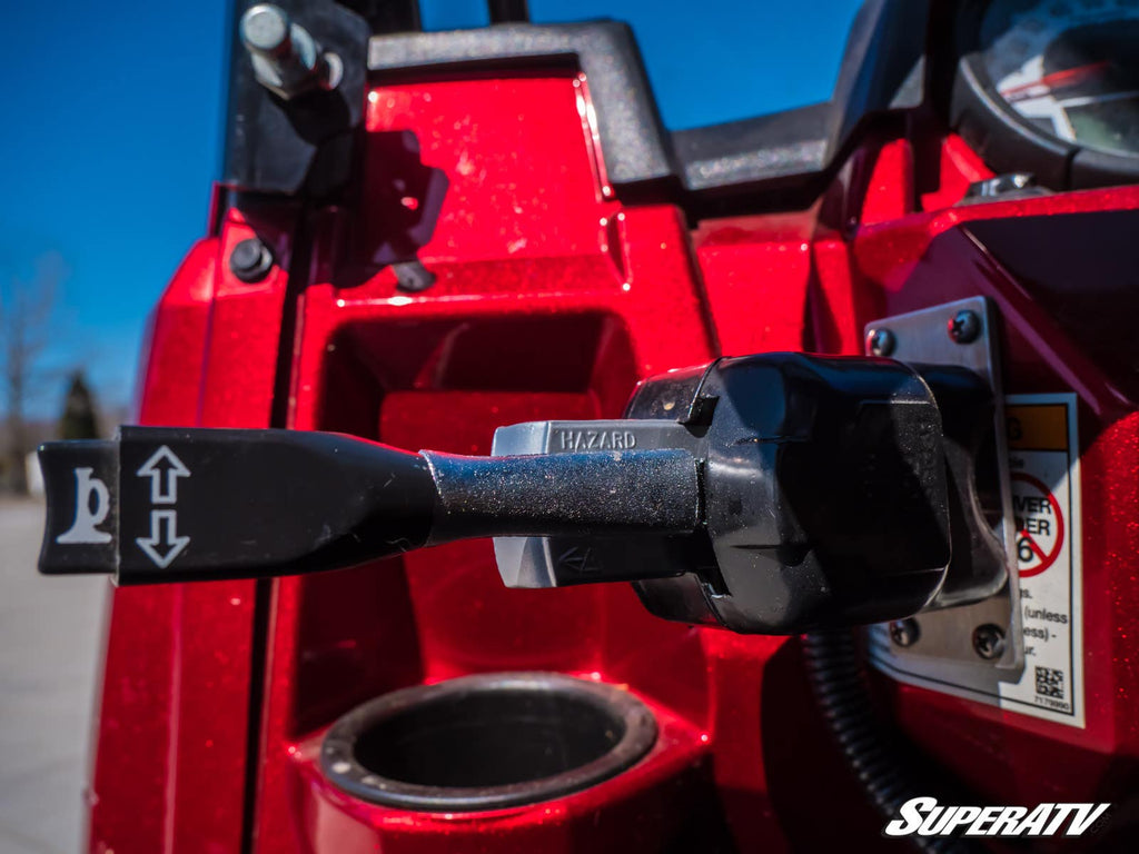 Close-up of a black lever labeled HAZARD on the bright red dashboard of a Polaris RZR RS1 featuring the Plug & Play Turn Signal Kit, with arrow symbols. A blurred blue sky and outdoors are in the background. The SuperATV logo is in the bottom right corner