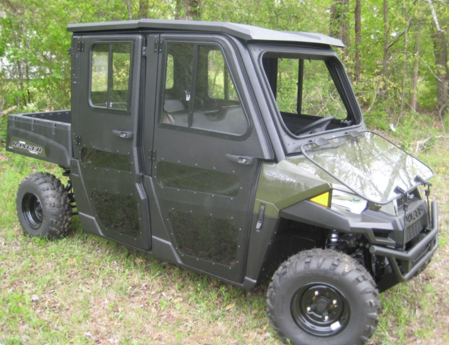 A Polaris Ranger Crew 1000 Armor Tech Roof UTV in dark green with four doors, black trim, rugged tires, a rear cargo bed, and large windows is parked on grass amid trees and greenery in a wooded outdoor area.