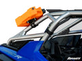 A Universal Jerry Can Cage Mount holds a bright orange fuel canister to the silver roll cage of a blue off-road vehicle above the passenger area. Black mounting hardware, interior details, and the “SuperATV” logo are also visible.