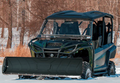A Rival Powersports Universal UTV Snow Plow Kit with Stamped Steel Blade (54/60/72 includes mount) is attached to a green and black off-road utility vehicle parked on snow, with leafless trees and dry grass in the background.