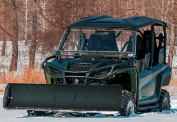 A Rival Powersports Universal UTV Snow Plow Kit with Stamped Steel Blade (54/60/72 includes mount) is attached to a green and black off-road utility vehicle parked on snow, with leafless trees and dry grass in the background.