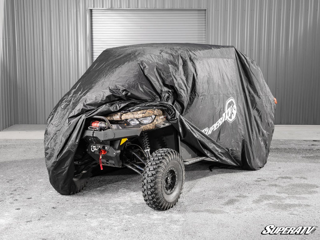 A UTV is partially covered by a black Universal UTV Cover. The rear, showing a large off-road tire, winch, and camo-patterned seat, is visible. The vehicle sits on concrete in front of a gray metal building with a closed garage door.