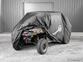 A UTV is partially covered by a black Universal UTV Cover. The rear, showing a large off-road tire, winch, and camo-patterned seat, is visible. The vehicle sits on concrete in front of a gray metal building with a closed garage door.