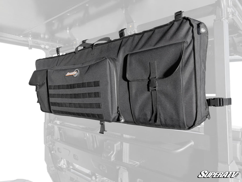 The UTV Gun Bag is a large black storage bag with zippered and flap pockets, MOLLE webbing, and a SuperATV logo patch on the main pocket. Its attached to a utility vehicles rear window frame against a mostly white background.