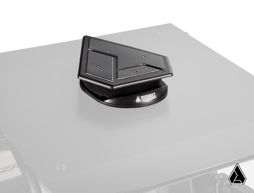 The Assault Industries UTV Roof Fan is a black, angular roof vent with a metallic finish and sharp lines, mounted on a flat gray surface. The base displays the “ASSAULT” logo, while a small triangular logo appears in the bottom right corner.