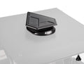 The Assault Industries UTV Roof Fan is a black, angular roof vent with a metallic finish and sharp lines, mounted on a flat gray surface. The base displays the “ASSAULT” logo, while a small triangular logo appears in the bottom right corner.