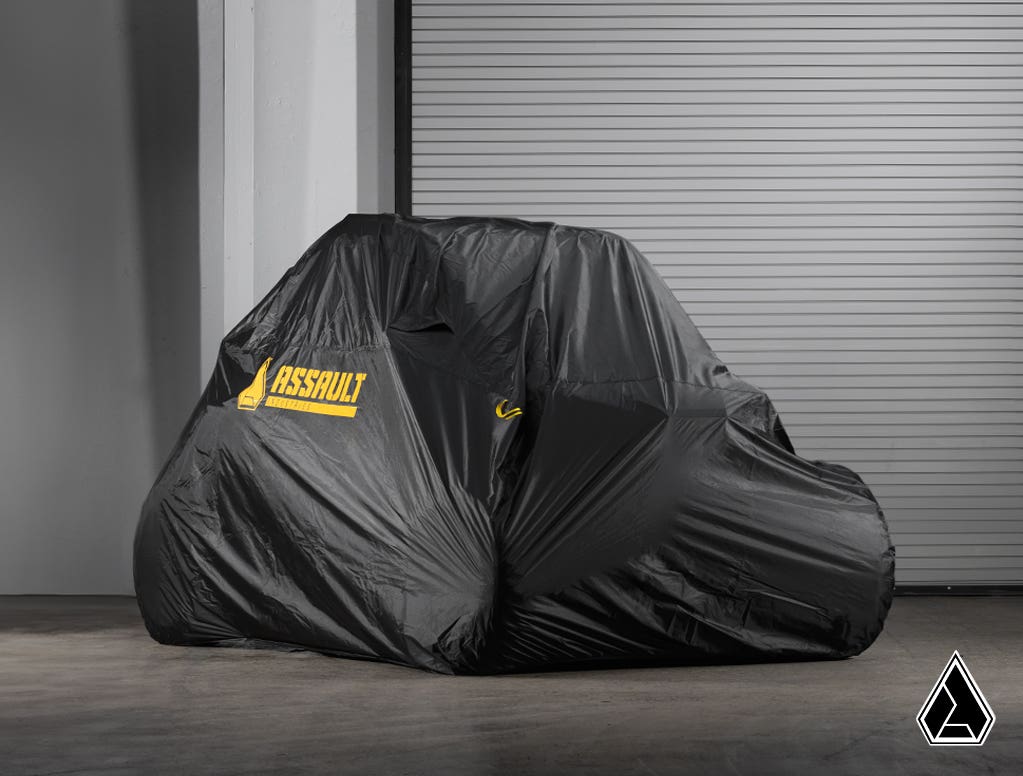 The Assault Industries Universal UTV Cover in black with a yellow ASSAULT logo conceals an off-road vehicle on a concrete floor before a gray metal roll-up door. A small triangular logo is visible in the lower right corner of the image.