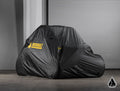 The Assault Industries Universal UTV Cover in black with a yellow ASSAULT logo conceals an off-road vehicle on a concrete floor before a gray metal roll-up door. A small triangular logo is visible in the lower right corner of the image.