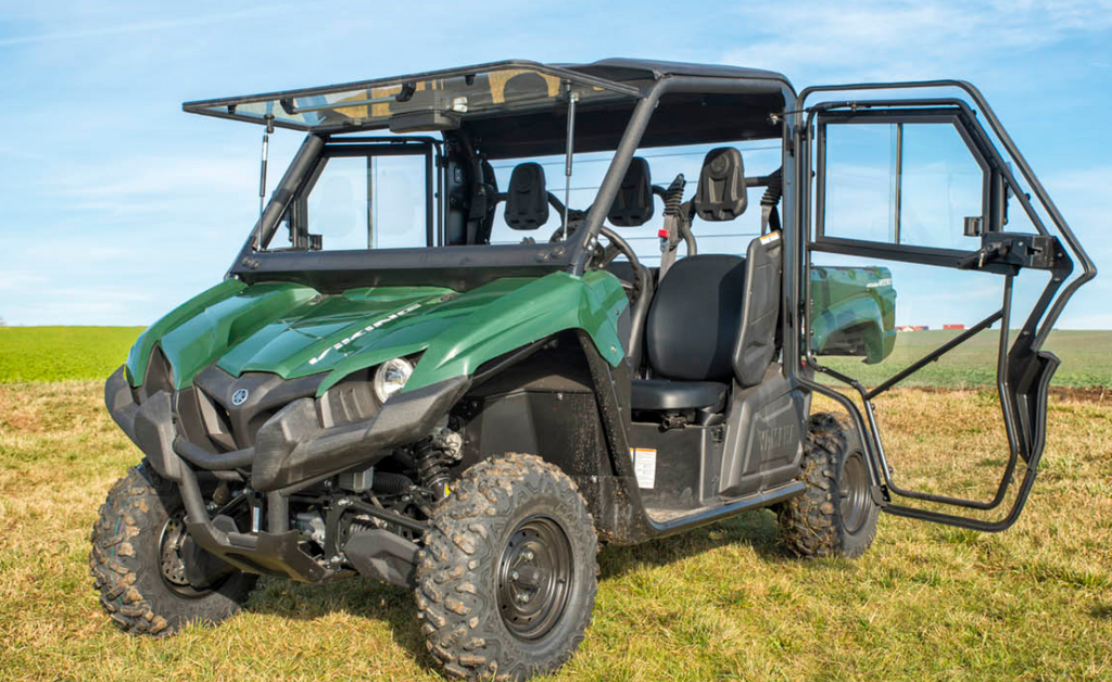 A Yamaha Viking fitted with a Hard Cab Enclosure featuring black roll bars and transparent doors sits on grassy terrain with its driver-side door open, showing two black seats. The backdrop includes blue skies and open fields.