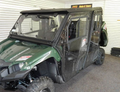 A Yamaha Viking VI equipped with a Hard Cab Enclosure featuring four transparent doors, rugged tires, and a black roof is parked indoors on carpeted flooring. Visible seating and white slatwall panels line the room’s walls.